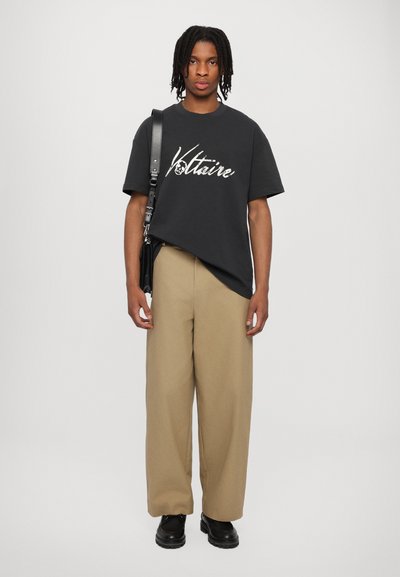 Schwarzes Baumwoll-T-Shirt mit dem Schriftzug "Voltaire" in kontrastierendem Weiß, kombiniert mit beigen weitem Hosenbein und schwarzen Schuhen.