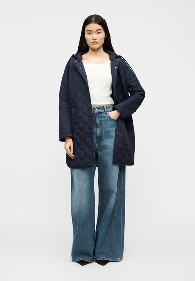 Γυναίκα που στέκεται φορώντας ένα ναυτικό μπλε καπιτονέ μπουφάν, λευκό τοπ με τετράγωνο ντεκολτέ και φαρδιά μπλε τζιν παντελόνια, μπροστά σε απλό ανοιχτό γκρι φόντο.