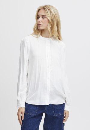 Weiße Bluse mit langen Ärmeln, einem Rüschenausschnitt, Knopfleiste und strukturiertem Stoff. Kombiniert mit blauen Jeans.