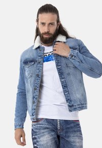 Hellblaue Denimjacke mit Sherpa-Kragen, ausgestattet mit silberfarbenen Knöpfen, Brusttaschen und tailliertem Schnitt. Wird über einem weißen Grafik-T-Shirt getragen.