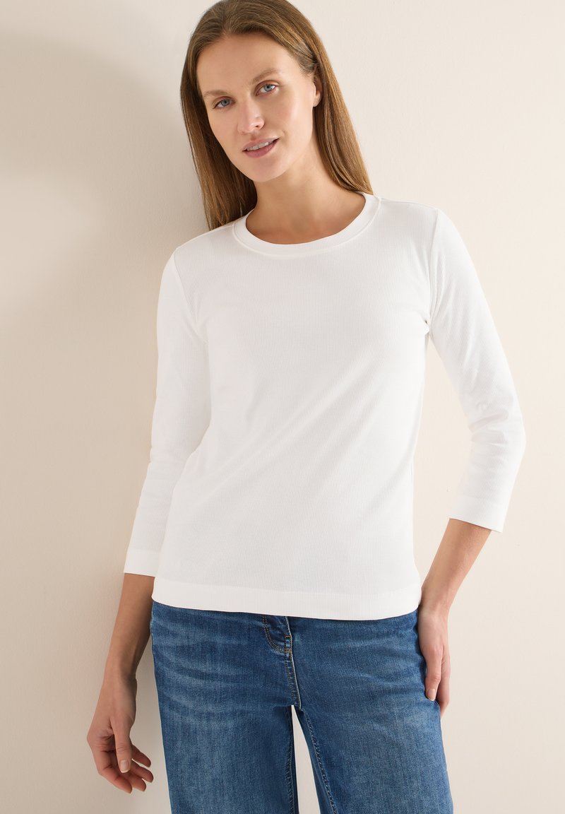 Haut blanc à manches longues côtelé avec un col rond, associé à un jean denim bleu. Les matériaux semblent doux et ajustés.