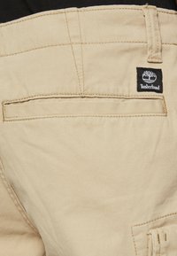 Béžové bavlněné cargo kalhoty s kapsou na zadní straně a černou nášivkou Timberland. Obsahují stehové detaily a odolnou tkaninu.