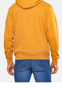 Felpa gialla con tasca a marsupio, polsini e orlo a coste. Abbinata a jeans blu con tasche posteriori e dettagli di cucitura.