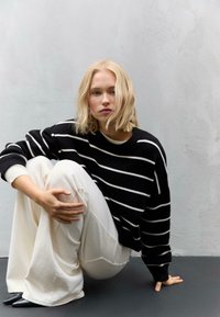 Maglione a righe bianche e nere con scollo a girocollo, abbinato a pantaloni bianchi larghi e scarpe nere a punta, su uno sfondo grigio texturizzato.