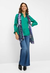 Grüne Satin-Bluse mit Knöpfen, kombiniert mit weiten blauen Jeans. Ein transparenter, gemusterter Schal umhüllt den Hals und zeigt lila und florale Details.