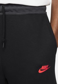 Nahaufnahme von schwarzen Nike-Sweatpants mit grauem Bündchen und rotem Nike-Logo Nähe der Tasche auf der rechten Seite.