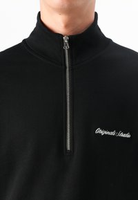 Et nærbillede af en person iført en sort zip-trøje med "Originals Studio" broderet på brystet. Baggrunden er helt hvid.