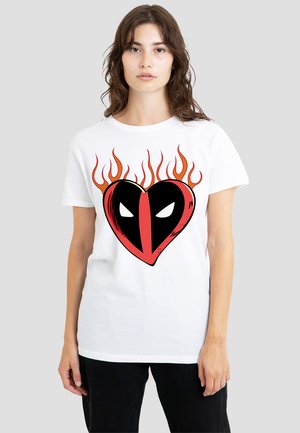 Woman wearing a white t-shirt with a flaming heart design resembling Deadpool’s mask in red and black on a gray background.