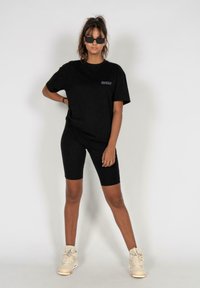 Sort oversized t-shirt med lilla logo, parret med sorte cykelshorts og cremegule høj-top sneakers, båret af en model.