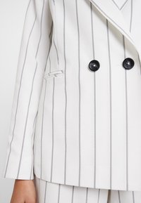 Valkoinen kaksoisnapitus bleiseri mustilla pystysuorilla raitoja, jossa on kaksi mustaa nappia, etutasku ja sileä kankaan rakenne.