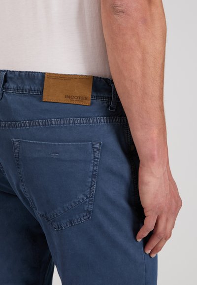 Man wearing blue casual pants with a brown leather brand label at the waistband, visible back pocket, and his relaxed right arm by his side.
