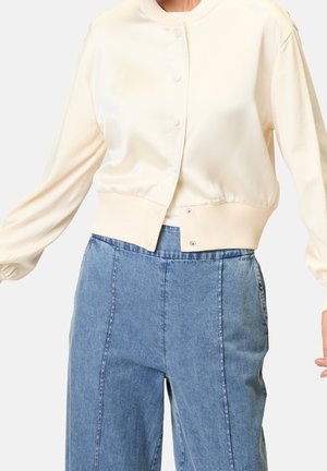 Personne portant un cardigan court crème boutonné à manches longues et un jean en denim bleu taille haute, bras légèrement étendus.