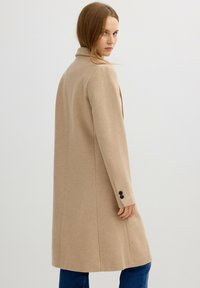 Abrigo de mezcla de lana beige con un diseño entallado, mangas largas y botones negros. Textura suave y corte recto, llegando hasta la mitad del muslo.