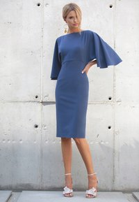 Granatowa dopasowana sukienka z rękawami w kształcie skrzydeł nietoperza, długości do kolan i gładkiej teksturze. Połączona z jasnoróżowymi sandałami z akcentami w kształcie kokardy.