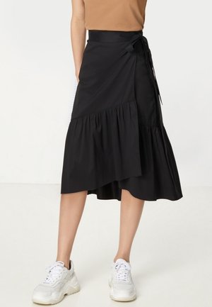 Femme portant une jupe portefeuille noire taille haute avec un détail à volants, associée à des baskets blanches épaisses et un haut marron sans manches.