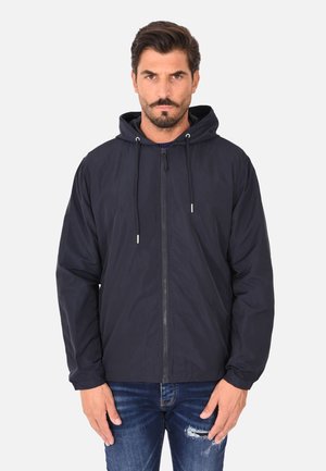 Uomo con capelli scuri e barba che indossa una giacca nera con cappuccio e zip, e jeans blu consumati, in piedi davanti a uno sfondo chiaro e uniforme.