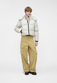 Hvit puffjakke med glidelås foran og avtagbar hette, kombinert med beige cargo bukser som har sidelommer og en avslappet passform. Sorte støvler.