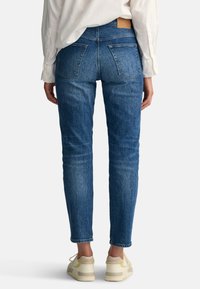 Slim-fit donkerblauwe denim jeans met een middenhoogte taille, vijf zakken en een tan leren label op de achterkant. Gemaakt met lichte sneakers.