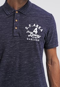 Marinblå polo-shirt med texturerad tyg, tre träknappar och vit tryckt text som säger "S.F. AREA 4 Fours Liners Oakland."