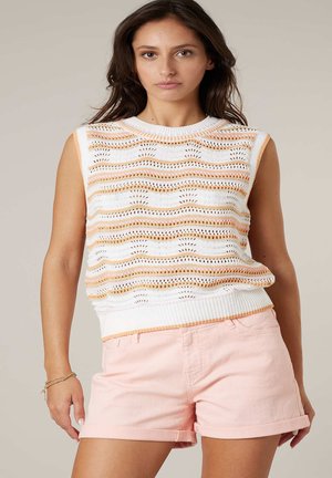 Femme portant un haut sans manches en maille blanche avec des rayures orange et un short rose clair à ourlet retroussé, debout devant un fond neutre.