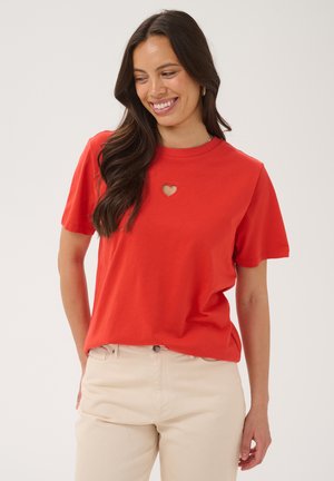 Femme souriante regardant vers le bas, portant un t-shirt rouge à manches courtes avec une petite découpe en forme de cœur près du col et un pantalon beige.