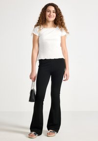 Jonge vrouw met krullend haar, gekleed in een witte top met korte mouwen, zwarte uitlopende broek, zilveren sandalen en een kleine zwarte handtas.