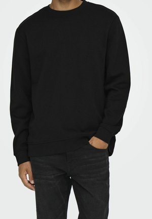 Mand iført sort sweatshirt med lange ærmer og mørke jeans, står med den ene hånd i lommen mod en ensfarvet baggrund.