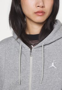 Harmaa vetoketjuhuppari, joka on valmistettu teksturoidusta kankaasta. Siinä on valkoinen Jumpman-logo vasemmassa rinnassa ja vaaleanharmaat nyörit hupussa.