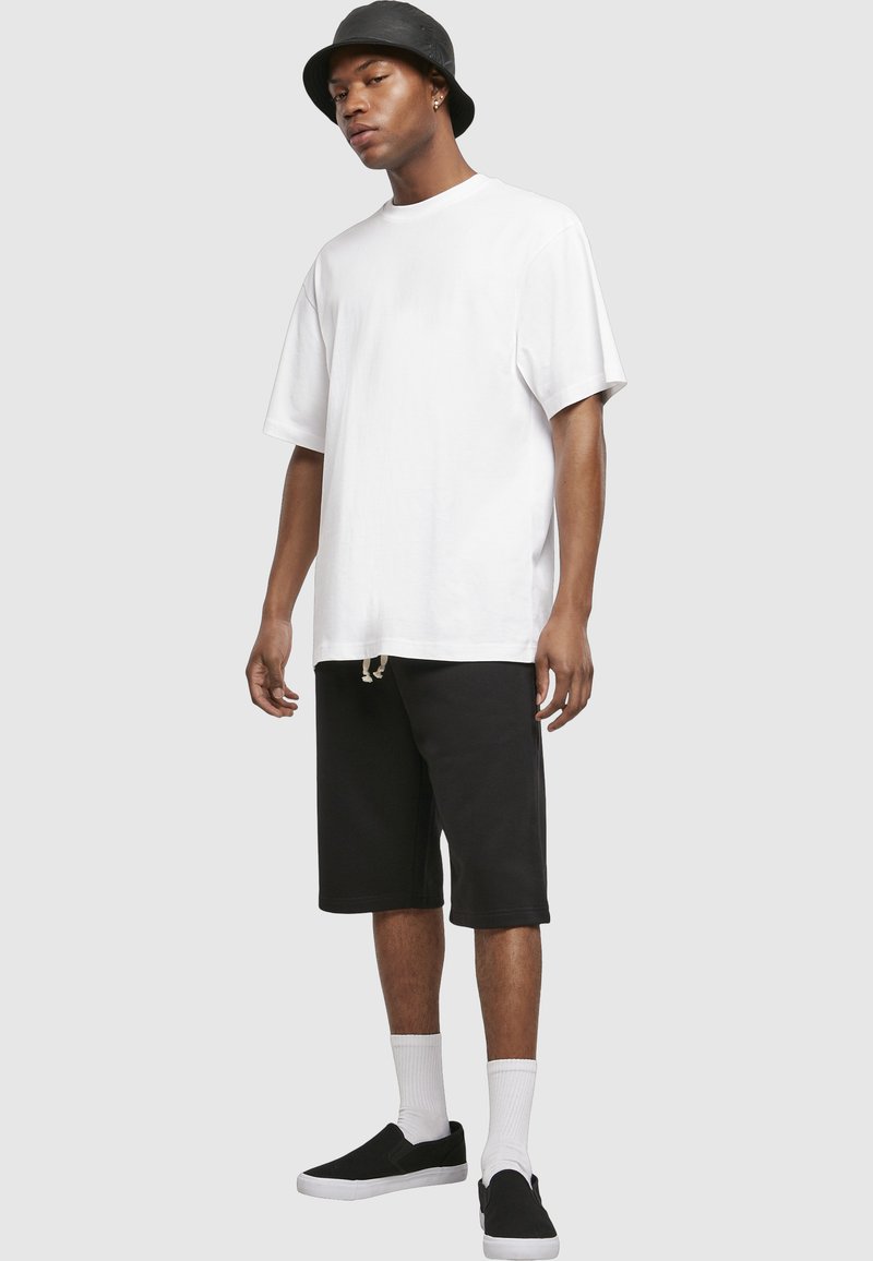 Mladi moški z črno kanti kapo, prevelikim belim majico, črnimi kratkimi hlačami do kolen, belimi nogavicami in črnimi natikači.