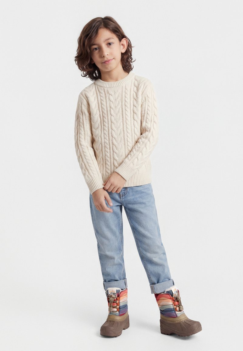 Enfant aux cheveux bruns mi-longs portant un pull crème à torsades, un jean bleu clair retroussé, et des bottes d'hiver colorées, debout devant un fond blanc.
