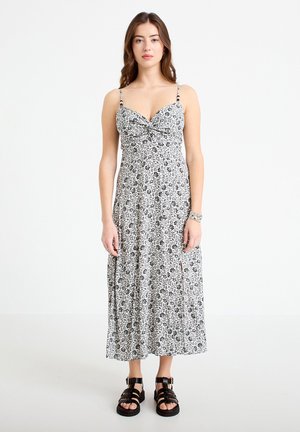 Młoda kobieta ubrana w czarno-białą sukienkę midi w kwiaty z cienkimi ramiączkami i czarne masywne sandały, stojąca na tle jednolitego białego tła.