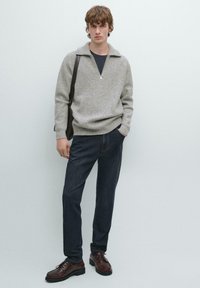 Pullover lavorato a maglia di un grigio chiaro con colletto mezza zip, abbinato a jeans blu scuro e scarpe stringate marroni. Il modello sta in piedi con una borsa a tracolla.