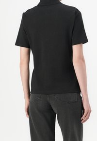 Personne portant un t-shirt noir uni à manches courtes et un jean gris foncé, vue de dos sur un fond blanc uni.