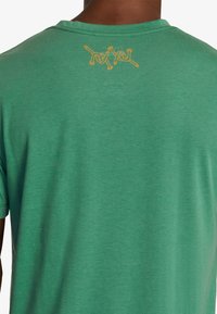 Green short-sleeve T-shirt featuring a rounded neckline and a graphic in orange on the upper back. Material appears soft and textured.