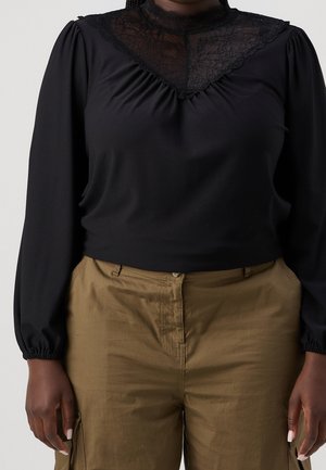 Personne portant un haut noir à manches longues avec un détail en dentelle au niveau du col et un pantalon marron taille haute, debout, les mains le long du corps.