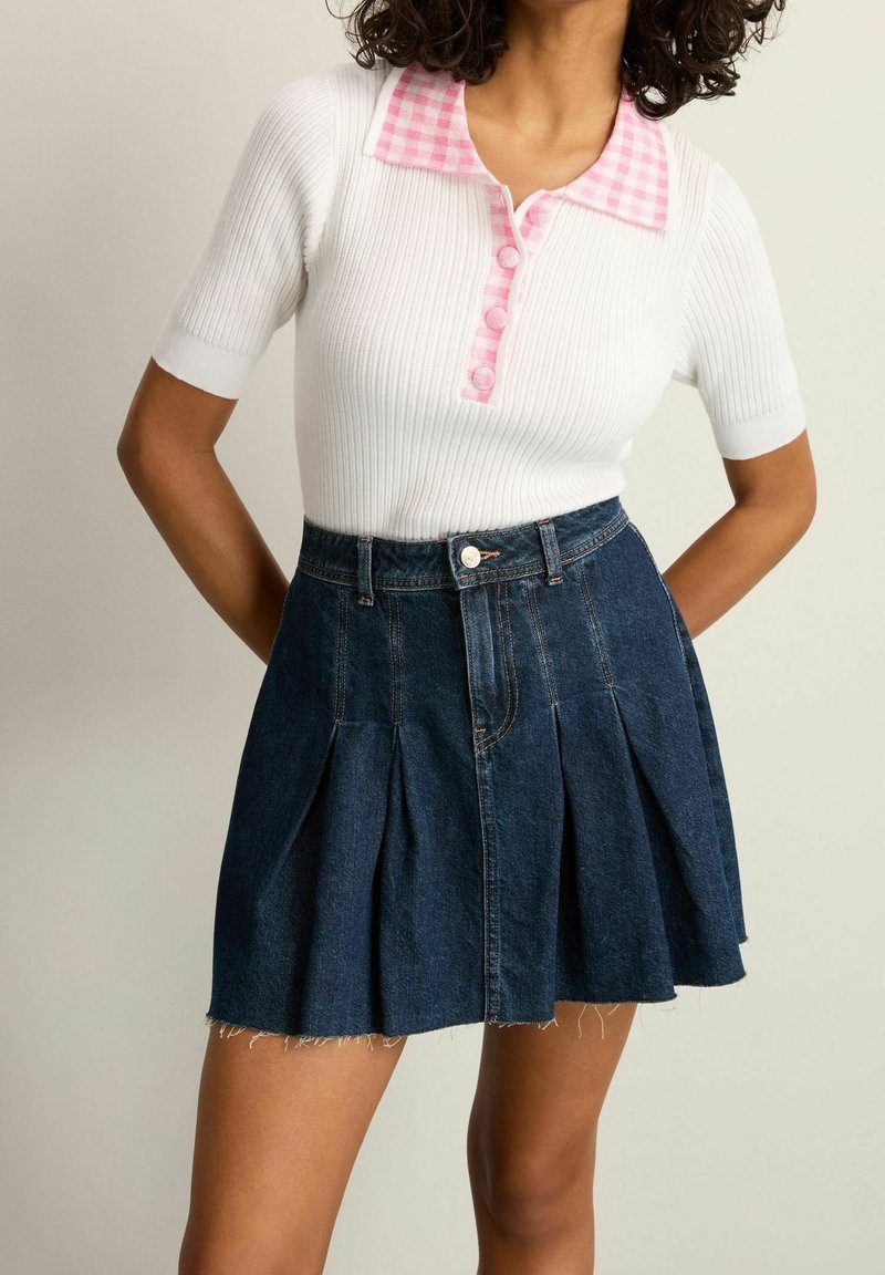Weißes geripptes Oberteil mit pinkem Karokragen und Knöpfen; kombiniert mit einem dunklen Jeansrock mit Falten und fransigem Saum. Lässige, figurbetonte Silhouette.
