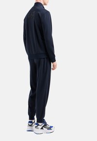 Tuta navy con pantaloni abbinati, realizzata in tessuto liscio. Presenta un logo sulla parte posteriore della giacca e scarpe sportive con dettagli blu.