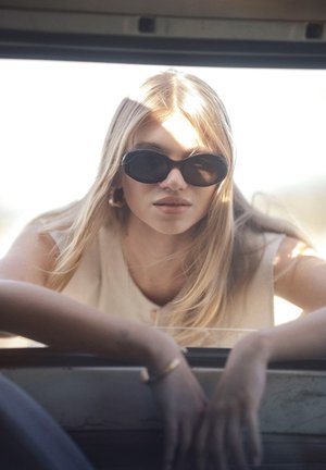 Blondynka w ciemnych okularach przeciwsłonecznych pochyla się do przodu, opierając ręce na ramie okna samochodu w jasnym świetle słonecznym.