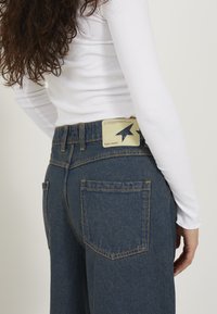 Pantalones de mezclilla de talle alto, color azul oscuro, con costuras en contraste amarillas. Presenta una etiqueta en la cinturilla trasera con un diseño de flecha.