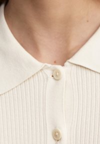 Krémová pletená blůza s ribbed vzorem, klasickým límcem a dvěma knoflíky na výstřihu. Měkká textura a jemný lesk.