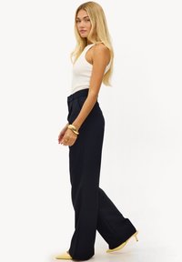 Débardeur blanc côtelé associé à un pantalon large taille haute de couleur bleu marine. Le mannequin porte des chaussures à talons jaunes et un bracelet en or.