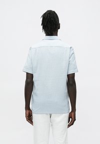 Korte mouw, lichtblauw overhemd met patroon, voorzien van een kraag, zachte textuur en subtiel ruitjesontwerp. Achteraanzicht toont een ontspannen pasvorm en naden.