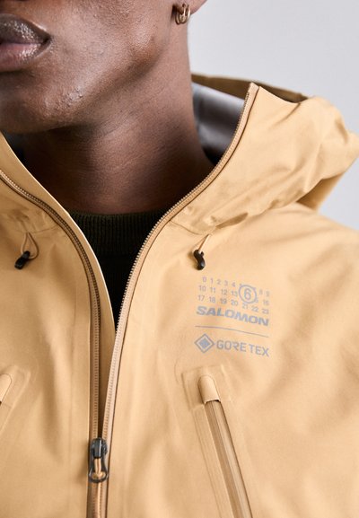 Veste GORE-TEX de couleur beige clair avec une fermeture éclair frontale, des poches latérales et un logo imprimé. Dispose d'une capuche réglable et d'une texture lisse et élégante.