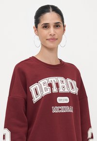Sweatshirt cor bordeaux com o texto "DETROIT" e "MICHIGAN" em branco, tecido suave, corte descontraído e costuras de ombro caídas.