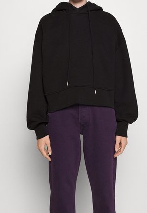 Personne portant un hoodie court noir avec des cordons et un pantalon violet taille haute, debout devant un fond uni.