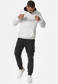 Sudadera blanca con embellecimientos negros y un bolsillo delantero, combinada con pantalones cargo negros y zapatillas blancas. La sudadera cuenta con una capucha de doble capa.