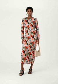 Mulher vestindo um vestido longo floral com padrões em vermelho, rosa e verde, segurando uma bolsa bege e usando sandálias de salto pretas.