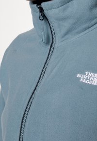 Polarowy zamknięty kurtka w delikatnym niebieskim kolorze. Posiada wysoki kołnierz, czarny zamek błyskawiczny oraz białe haftowane logo na piersi. Teksturowany, przytulny materiał.