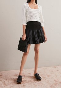 Vrouw in witte oogjesblouse, zwarte gelaagde rok, zwarte klompen en met een zwart franjerugzak in de hand, staand op een beige tapijt.