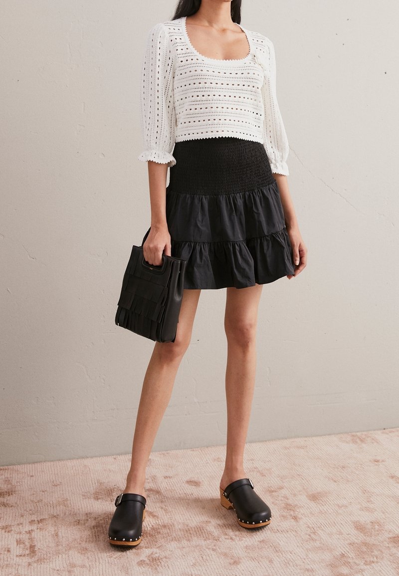 Vrouw in witte oogjesblouse, zwarte gelaagde rok, zwarte klompen en met een zwart franjerugzak in de hand, staand op een beige tapijt.